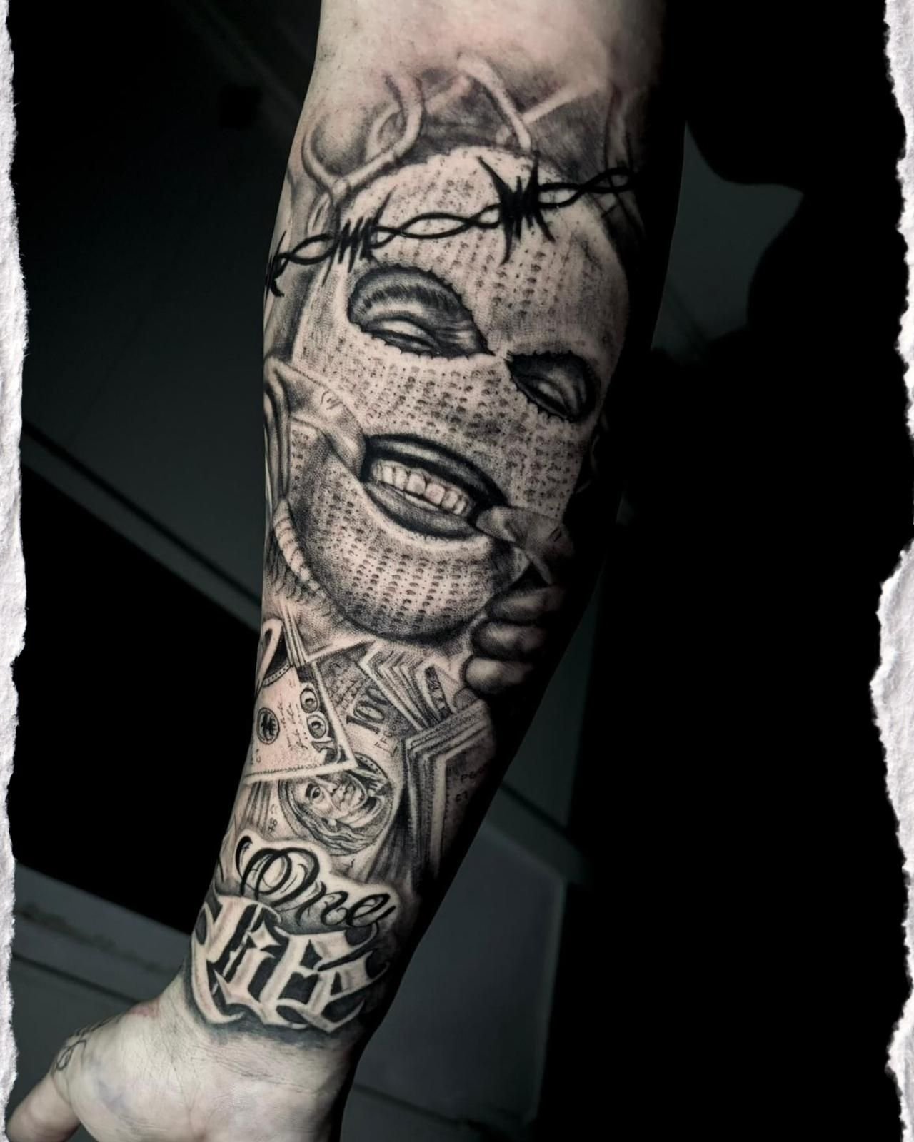 Antebrazo tatuado con rostro en blanco y negro y diseños de alambre de púas sobre un fondo negro.