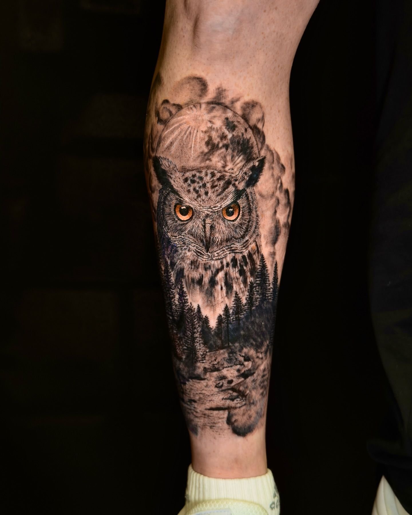 Tatuaje de un búho negro en la parte inferior de la pierna, con ojos naranjas y plumas detalladas.