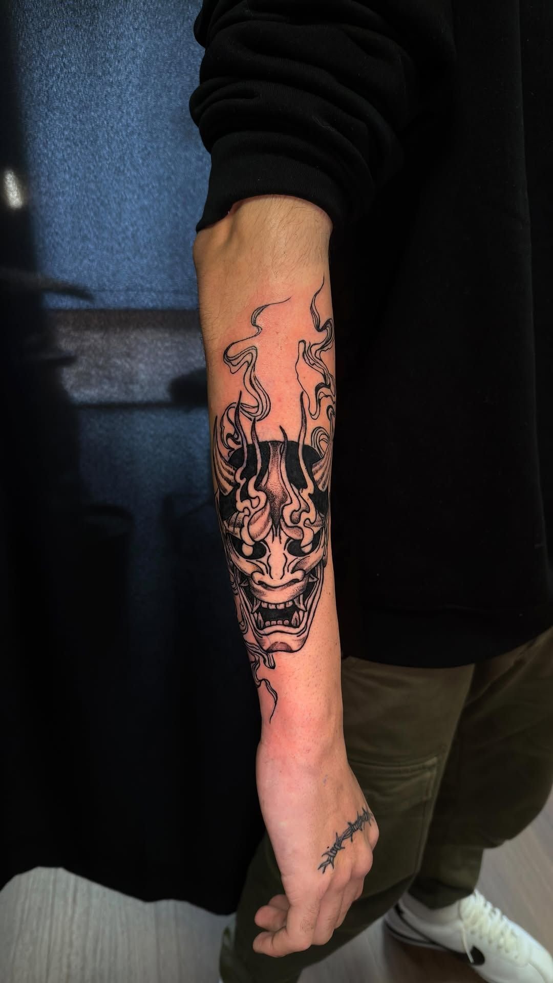 Brazo con un gran tatuaje negro de una calavera y un diseño floral, que cuelga junto a ropa oscura y pantalones verde oliva.
