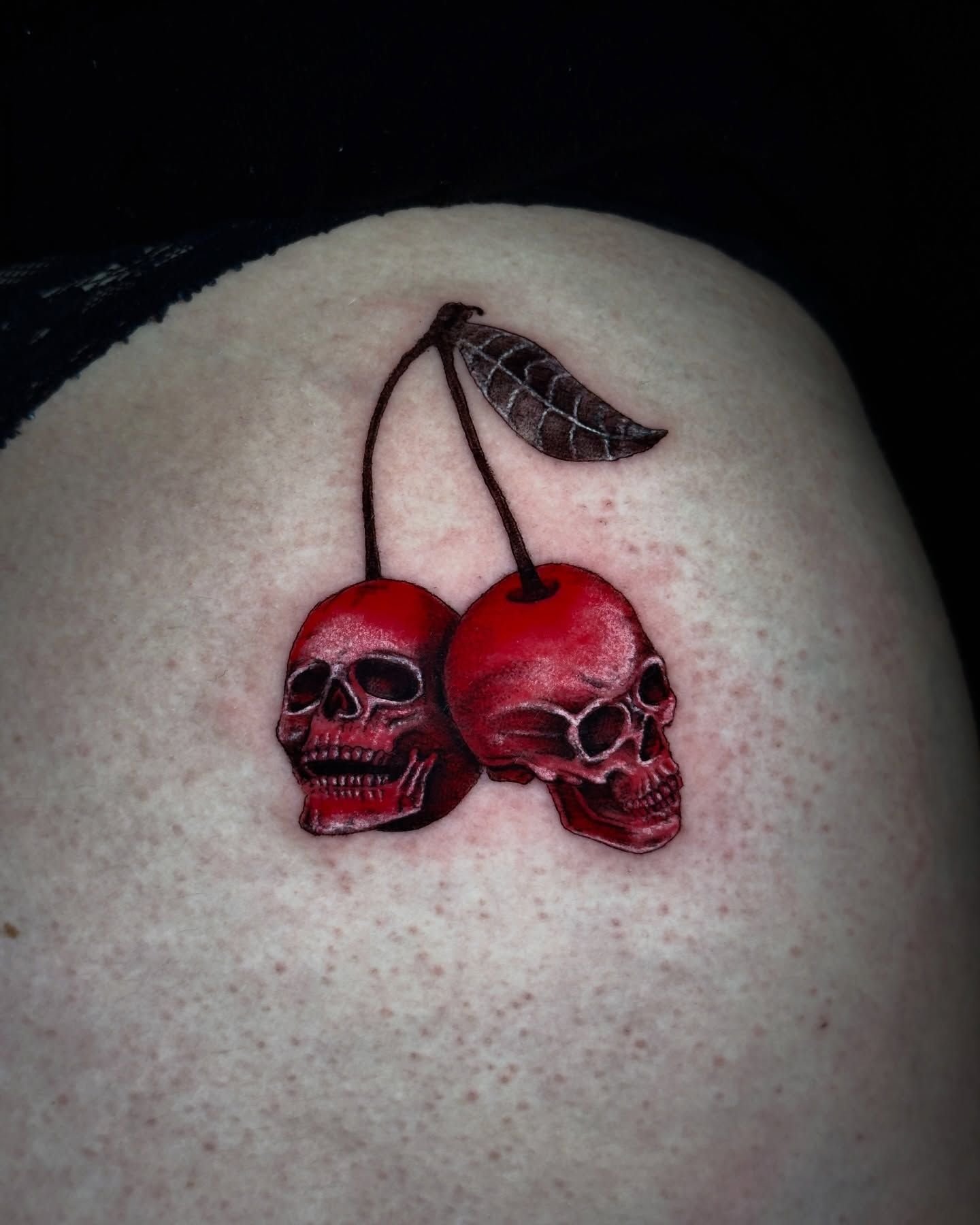 Tatuaje rojo de dos cerezas con forma de calaveras con una hoja en la piel.